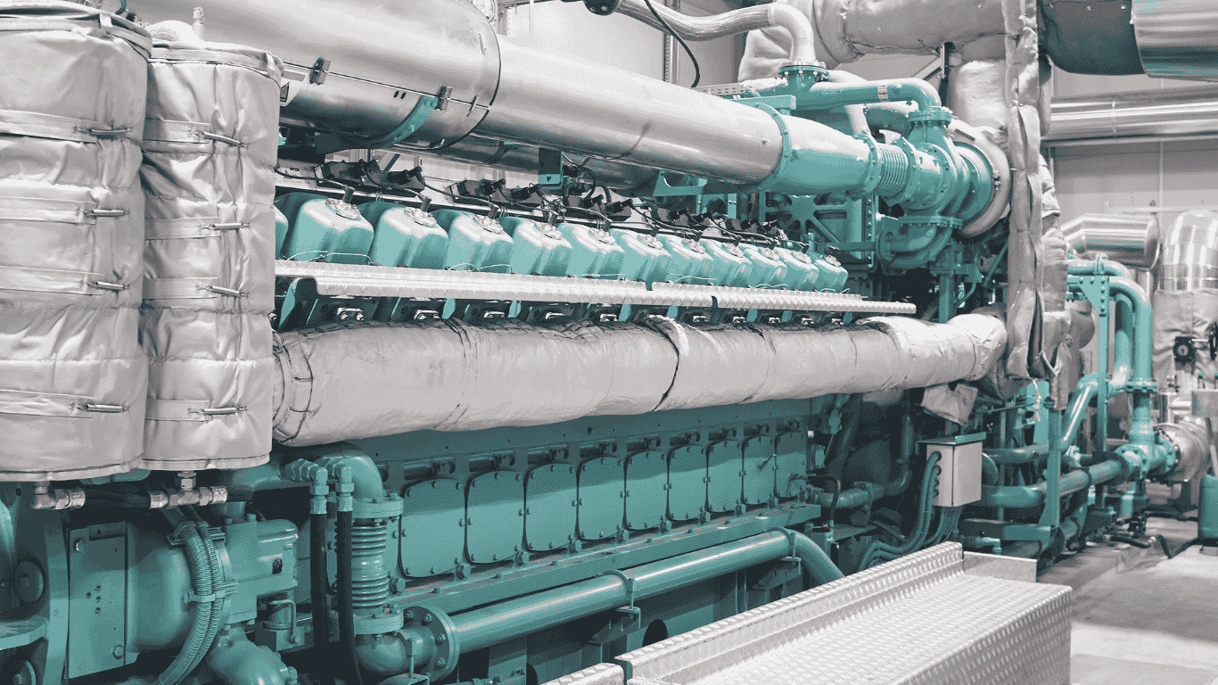 The image shows a large industrial engine or turbine with extensive piping, insulation, and heat recovery hardware, which is typical of a cogen plant where fuel is used to generate electricity and the waste heat is captured as steam for heating or process use. These systems are commonly installed to supply steam and power efficiently in facilities like hospitals, industrial sites, or a campus energy center serving multiple buildings across a campus.