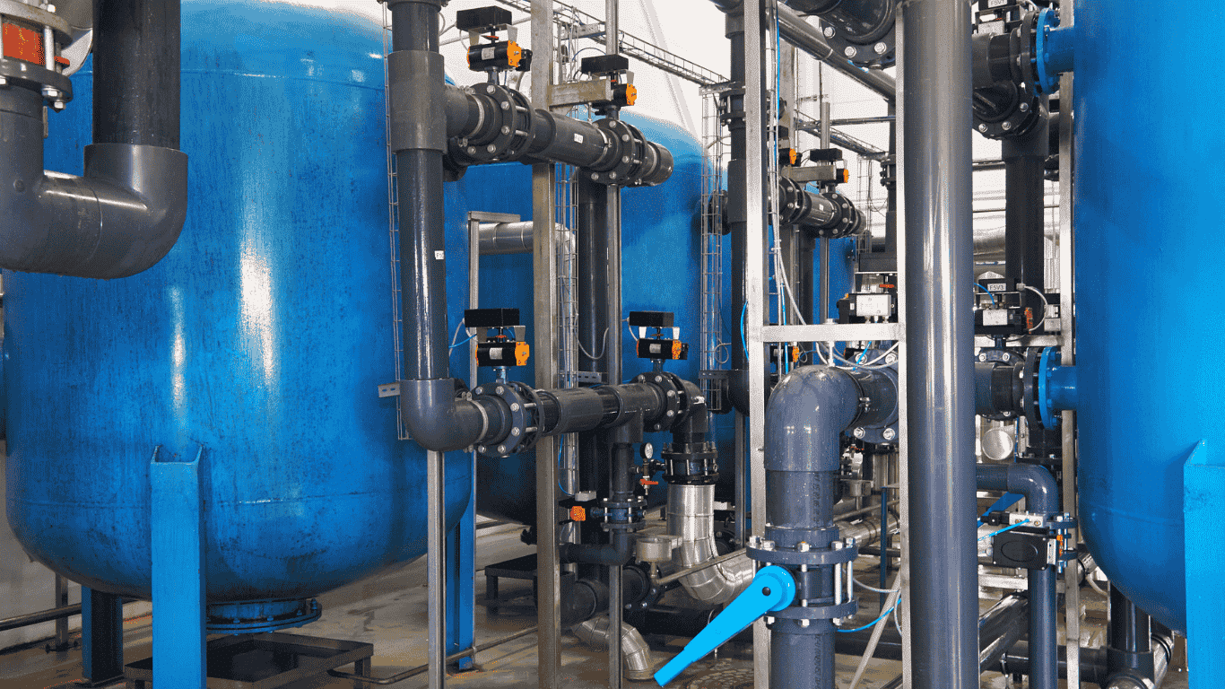 Industrial water softener system with tanks and piping designed to treat hard water, reduce water hardness, and prevent buildup like calcium carbonate and iron that can damage pipes. The system uses a resin bed filled with resin beads to remove grains of hardness, helping a household or facility determine what size and choose the right size water softener based on water hardness level, daily water usage, and water pressure.