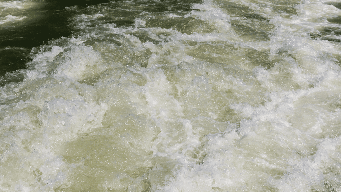 Close-up image of turbulent water used to illustrate turbidity sensors and real-time turbidity measurements in industrial and environmental monitoring systems. The moving water contains visible suspended particles, solid particles, and particulate matter that affect water quality, water clarity, and turbidity levels in a water body and natural waters. Modern turbidity instruments and nephelometric sensors use a light beam from an led lamp to emit light through a water sample, where light scattered by particles in the water is detected by a light detector or two light detectors. The amount of light, amount of light scattered, and how light energy scatters help measure turbidity, determine turbidity values, and generate a precise turbidity reading or final turbidity reading in nephelometric turbidity units and formazin nephelometric units for drinking water, food and beverage, and other industrial processes.