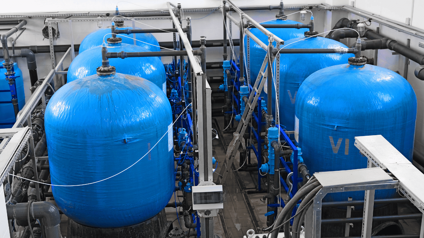 Industrial water softener system with multiple blue FRP pressure vessels and interconnected piping designed to remove hardness from hard water through ion exchange. The resin tank contains a resin bed where calcium and magnesium minerals are exchanged with sodium, producing soft water and softened water. The process improves water efficiency, supports reliable performance, and is commonly used for commercial and industrial water treatment.