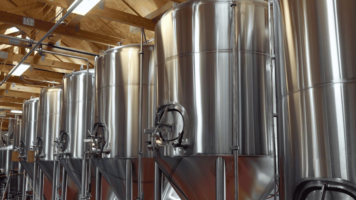 Stainless steel fermentation tanks inside a modern brewery in the food and beverage industry, showing the brewing process where beer is produced. The image represents a brewery wastewater system, process waste, spent grain, animal feed by-products, and production volumes that generate high strength wastewater, high strength effluents, biological oxygen demand, biochemical oxygen demand, oxygen demand BOD, total suspended solids, organic material, suspended solids, high BOD, and other industrial wastewater requiring wastewater treatment.